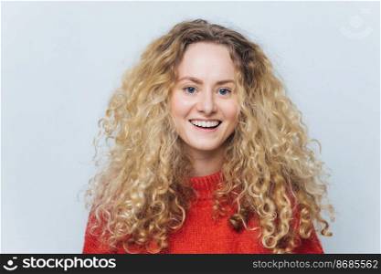 Isolated shot of attractive glad blonde gorgeous female has curly hair, has broad smile, being in high spirit after successful day, poses against blank white studio background. Delighted young woman