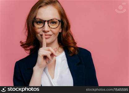 Isolated shot of attractive secret woman keeps fore finger over lips, demonstrates shush gesture, tells secret information, has healthy skin, manicure, makeup, wears formalwear, poses over pink wall