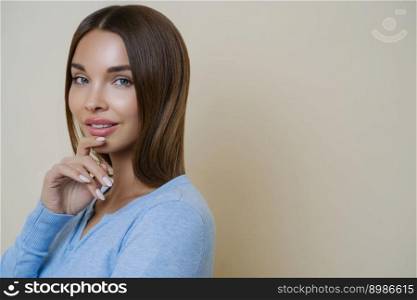 Isolated shot of attractive young beauty model touches chin gently, has manicure, looks at camera with blue eyes, considers future plans, p&ers her skin. Beige background with empty copy space