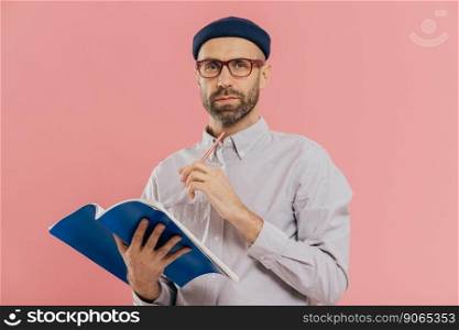 Isolated shot of attractive young male holds textbook, makes notes in organizer, dressed formally. Clever student prepares for examination or does home assignment, isolated over pink studio wall
