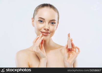 Isolated shot of beautiful girl applies lipstick on lips, has perfect makeup, poses with beauty product, touches face gently, poses shirtless, gets ready for work, poses against white background