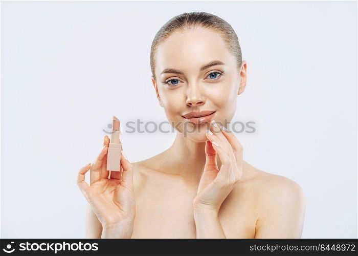 Isolated shot of beautiful girl applies lipstick on lips, has perfect makeup, poses with beauty product, touches face gently, poses shirtless, gets ready for work, poses against white background