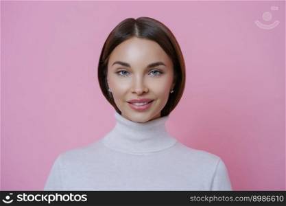 Isolated shot of charming glad young woman with pleasant smile, wears white turtleneck, looks directly at camera, isolated on pink background. Beautiful female student returns home from lectures