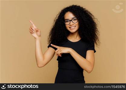 Isolated shot of happy African American woman points up and aside, demonstrates blank copy space for your advertisement, wears spectacles, casual t shirt, has gentle smile on face. People and promo
