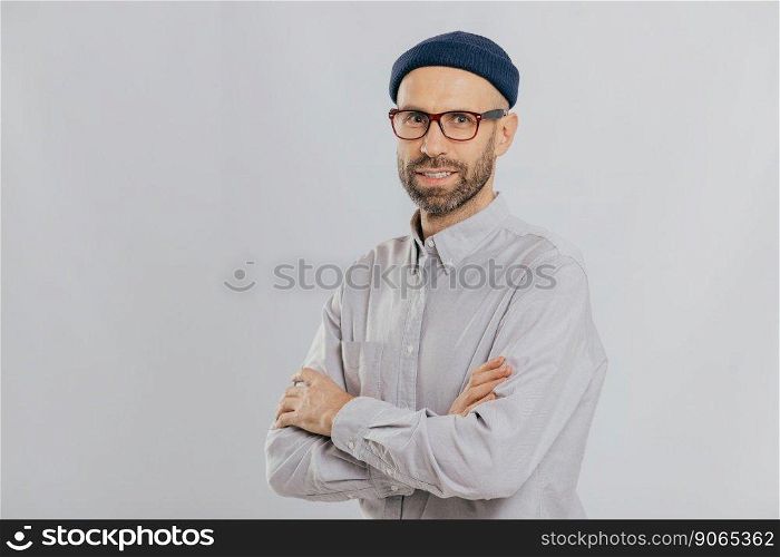 Isolated shot of pleasant looking unshaven male keeps hands crossed, wears stylish hat, optical glasses, models over white background with copy space for your advertising content, enjoys leisure