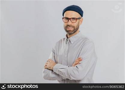 Isolated shot of pleasant looking unshaven male keeps hands crossed, wears stylish hat, optical glasses, models over white background with copy space for your advertising content, enjoys leisure
