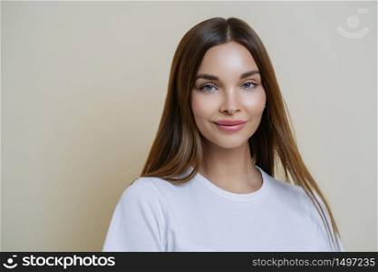 Isolated shot of pleasant looking woman with dark straight hair, wears white t shirt, isolated over brown background, copy space for your advertising content, listens interesting stories and good news