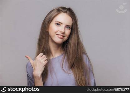 Isolated shot of pleased attractive brunette woman dressed casually, has happy expression, isolated over grey background with copy space for your advertisment or promotional text indicates with finger