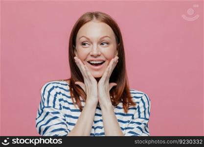 Isolated shot of pretty beautiful woman looks aside, keeps both hands on cheeks, dressed in striped casual sweater, demonstrates natural beauty, looks aside, isolated over pink studio background