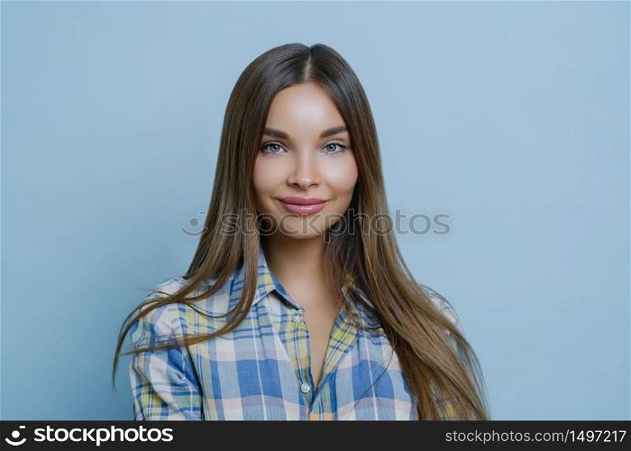 Isolated shot of pretty businesswoman has pleased face expression, tender smile, dressed in casual shirt, has clear skin and attractive facial features, gazes sensually at camera, blue background