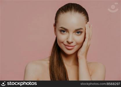 Isolated shot of pretty woman with combed pony tail, enjoys freshness of skin, looks with charming expression, stands bare shoulders, isolated on pink wall. Health care, makeup, spa and face lifting