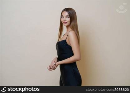 Isolated shot of pretty young European woman in black fashion dress, stands sideways, wears manicure and makeup, isolated over beige background. Fashion photo. Female model stands in profile