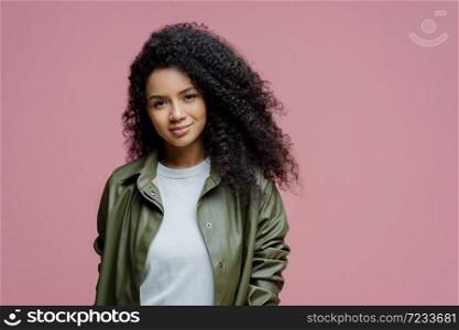 Isolated shot of serious dark skinned woman looks with confident calm expression at camera, listens something very attentively, dressed in stylish outfit, poses against pink studio background