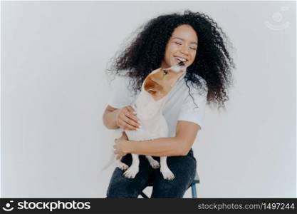 Isolated shot of smiling Afro American woman with curly hairstyle, gets kiss from favourite dog, sits at chair against white wall, wears t shirt and jeans. People, animals, emotions and love concept