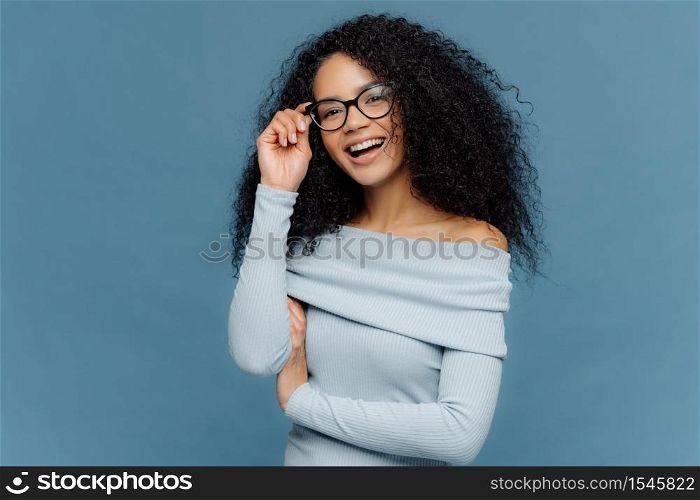 Isolated shot of smiling young African American woman touches frame of glasses, has glad expression, wears stylish jumper, isolated on blue background, happy spend free time with friends. Emotions