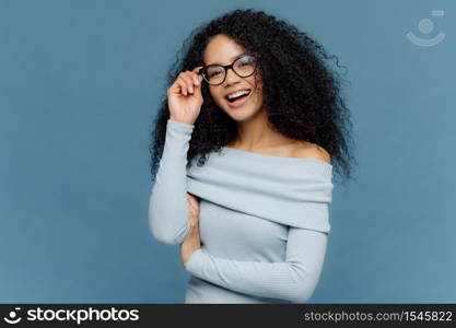 Isolated shot of smiling young African American woman touches frame of glasses, has glad expression, wears stylish jumper, isolated on blue background, happy spend free time with friends. Emotions