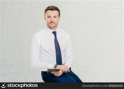 Isolated shot of successful male manager wears elegant white shirt, black trousers and tie, uses modern tablet computer for work and entertainement, isolated over white background. Business concept