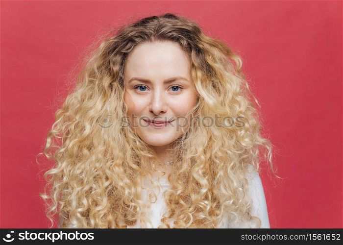 Isolated shot of young adorable female with curly hair and blue eyes, has glad expression, has appealing appearance, being in high spirit after good walk outside, poses against pink background