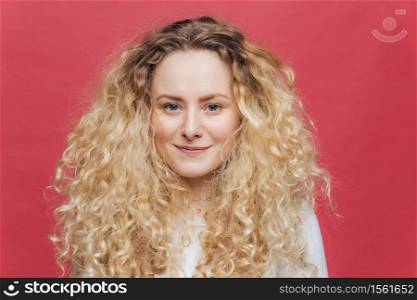 Isolated shot of young adorable female with curly hair and blue eyes, has glad expression, has appealing appearance, being in high spirit after good walk outside, poses against pink background