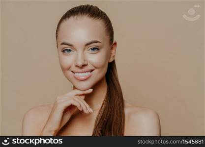 Isolated shot of young woman with European appearance has pure healthy smooth skin after doing daily cleansing procedures, smiles toothily, stands with bare shoulders indoor over beige wall.