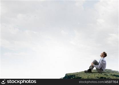 Isolated to find inspiration. Young smiling businessman sitting alone on rock top