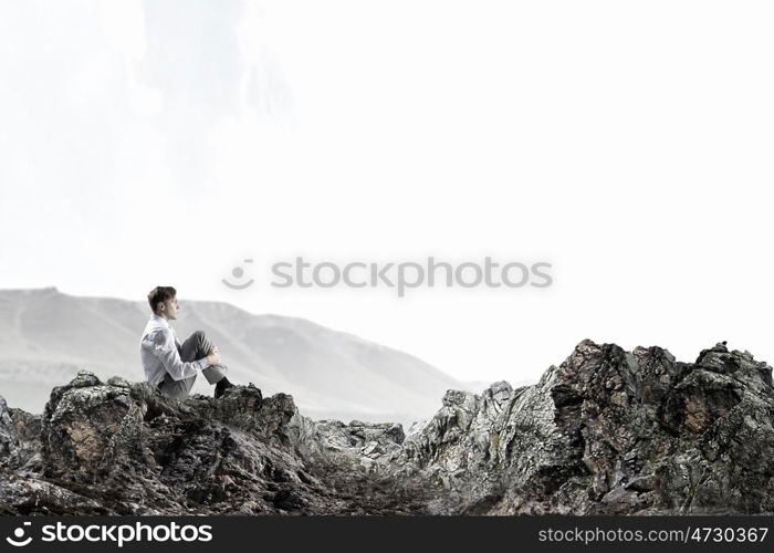 Isolated to find inspiration. Young thoughtful businessman sitting alone on rock top