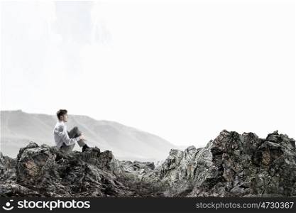Isolated to find inspiration. Young thoughtful businessman sitting alone on rock top