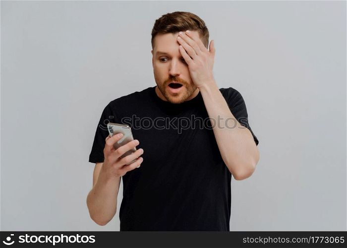 It can not be true. Young impressed unshaved ginger man reading latest news in internet on mobile phone, being in shock while receiving shocking message on smartphone, isolated over grey background. Impressed guy reading unexpected news in social media on smartphone with amazed expression
