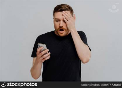 It can not be true. Young impressed unshaved ginger man reading latest news in internet on mobile phone, being in shock while receiving shocking message on smartphone, isolated over grey background. Impressed guy reading unexpected news in social media on smartphone with amazed expression