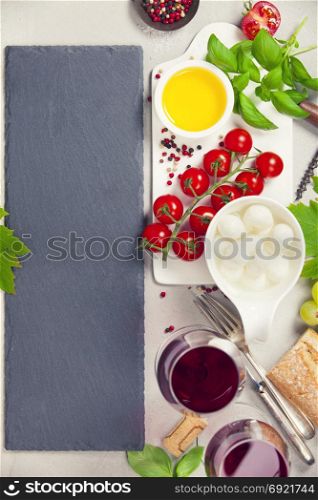 Italian antipasti snack for wine. Mozzarella cheese, fresh basil leaves, tomatoes, olive oil and glasses of red wine on concrete background, top view. Caprese salat ingredients