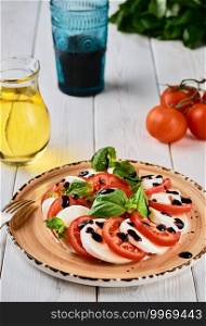 Italian Caprese salad with tomatoes, mozzarella cheese, basil balsamic vinegar and olive oil. On a ceramic plate on a white wooden table. Country style. Vegetarian dish