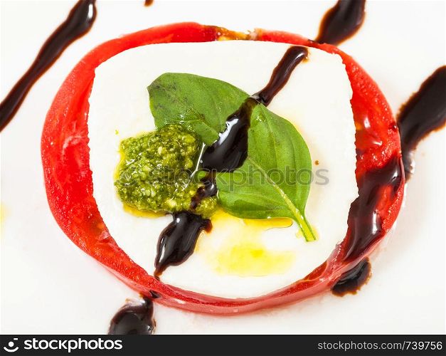 italian cuisine insalata caprese (caprese salad) - top view of stack from mozzarella cheese and tomato slices with basil leaf close up seasoned by olive oil, balsamic vinegar and pesto sauce on plate