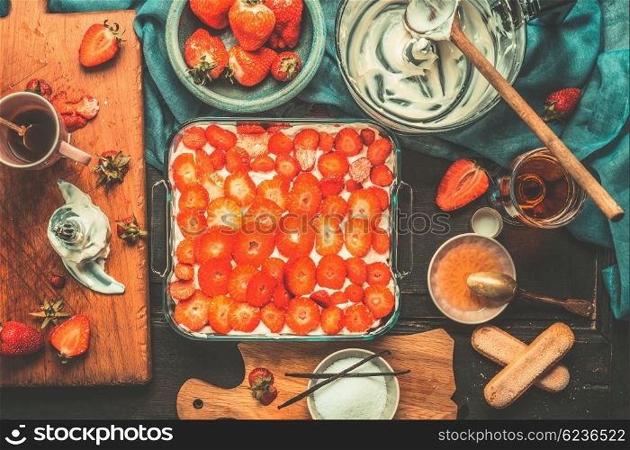 Italian Cuisine, strawberry tiramisu cake preparation with cooking ingredients and kitchen tools on dark vintage background, top view. Italian food concept