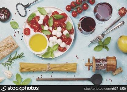 Italian food background. Mozzarella cheese, fresh basil leaves, tomatoes, olive oil, spaghetti and glasses of red wine on concrete background, top view. Caprese salat ingredients