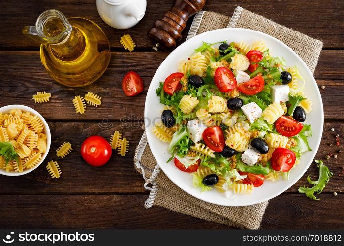 Italian pasta salad with fresh tomato, cheese, lettuce and olives on wooden background. Mediterranean cuisine. Cooking lunch. Healthy diet food