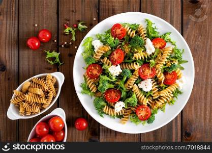 Italian pasta salad with wholegrain fusilli, fresh tomato, cheese, lettuce and broccoli on wooden rustic background. Mediterranean cuisine. Cooking lunch. Healthy diet food. Top view