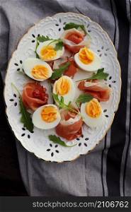 Italian salad. Boiled eggs with smoked ham and arugula on white plate. Dark wooden background