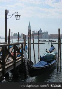 Italian traditional gondola parking near old wooden berth on the background of view of San Giorgio Maggior Church Venice, Italy.. Old wooden pier with parking italian tradition gondola.