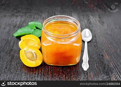 Jam apricot in glass jar, fruits, a spoon and a branch with green leaves on the background of dark wooden board