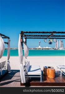 JAN 31, 2015 Abu Dhabu, UAE - Modern Beach Gazebo white curtain with white seat and wooden stool table. Blue sea under clear summer sky at Corniche beach with Marina island in background