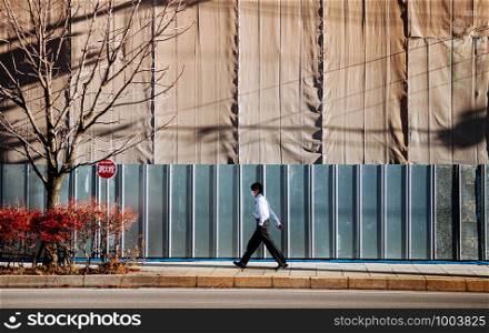 Japanese business man walking on sidewalk next to consturction site under bright winter sunlight