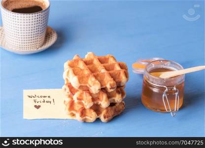 Jar of honey, fresh waffles and hot cup of coffee and a piece of paper with the message welcome Friday and a heart, on a blue wooden background.