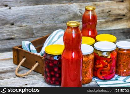Jars with variety of canned vegetables and fruits, jars with zacusca and bottles with tomatoes sauce. Preserved food concept in a rustic composition.