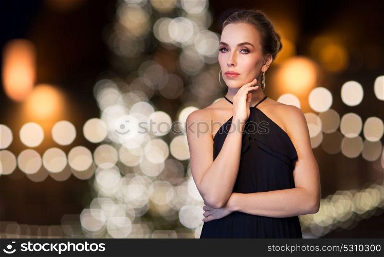 jewelry, holidays, luxury and people concept - beautiful woman in black wearing diamond earrings over christmas tree lights background. woman wearing jewelry over christmas lights