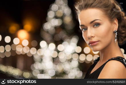 jewelry, holidays, luxury and people concept - beautiful woman in black wearing earrings over christmas tree lights background. woman wearing jewelry over christmas lights