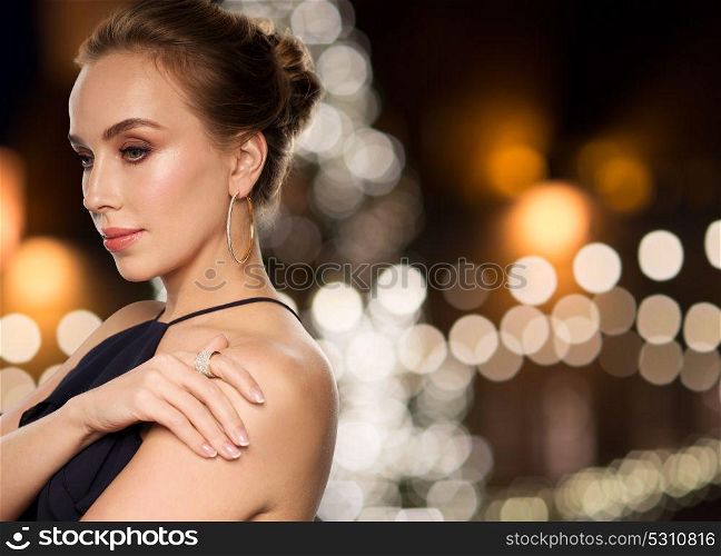jewelry, holidays, luxury and people concept - beautiful woman in black wearing diamond earrings over christmas tree lights background. woman wearing jewelry over christmas lights