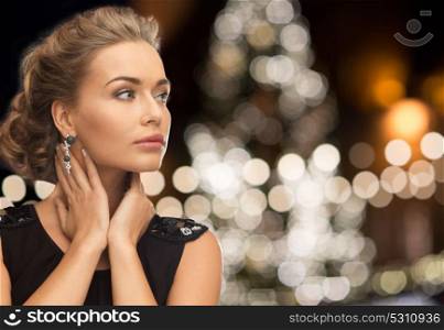 jewelry, holidays, luxury and people concept - beautiful woman in black wearing earrings over christmas tree lights background. woman wearing jewelry over christmas lights