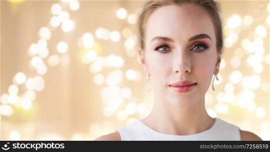 jewelry, luxury and people concept - portrait of woman in white dress with diamond earring over beige background and festive lights. woman in white dress with diamond earring