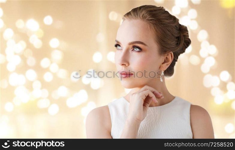 jewelry, luxury and people concept - portrait of woman in white dress with diamond earring over beige background and festive lights. woman in white dress with diamond earring