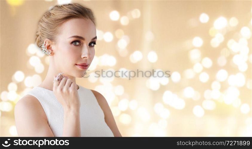 jewelry, luxury and people concept - portrait of woman in white dress with diamond earring and finger ring over beige background and festive lights. woman in white dress with diamond jewelry